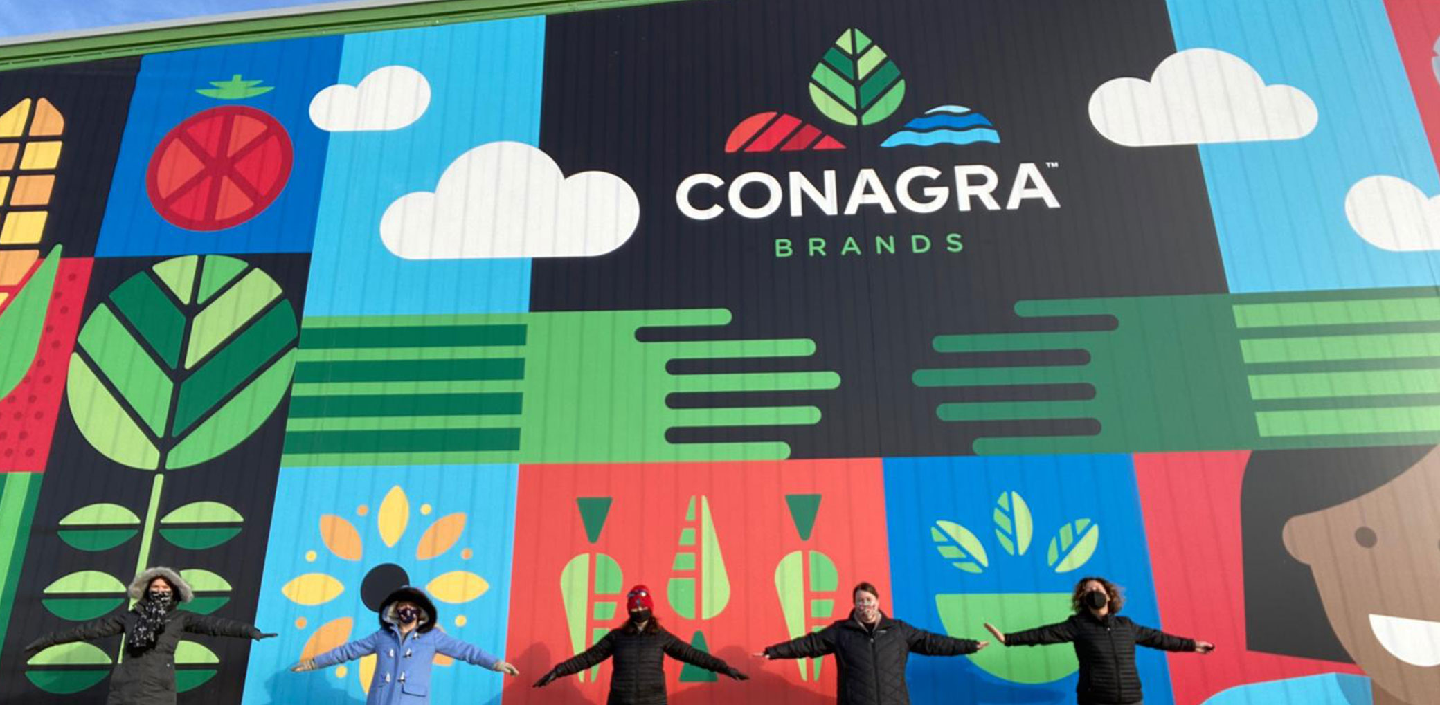 Five people with outstretched arms standing in front of a graphic, colorful Conagra mural